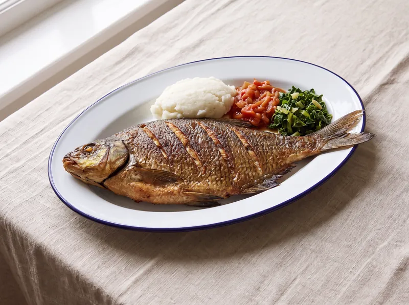 A whole cooked bream on an enamel plate with pap, tomato relish and greens